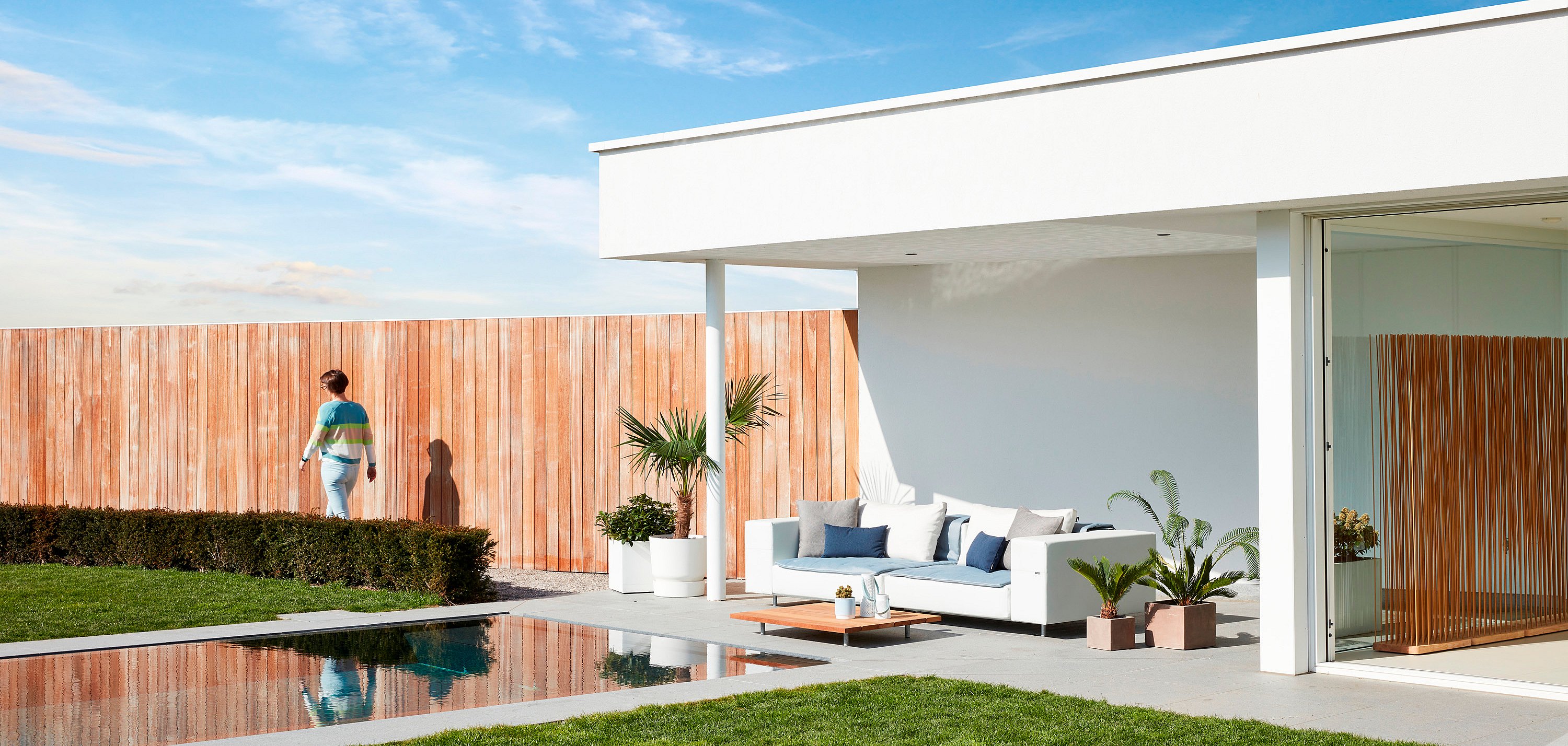 Weißes Walross-Set im Freien, das in der Abenddämmerung auf einer Holzterrasse neben einem Pool beleuchtet wird und sanftes Licht auf eine geschwungene hölzerne Gartenmauer wirft.