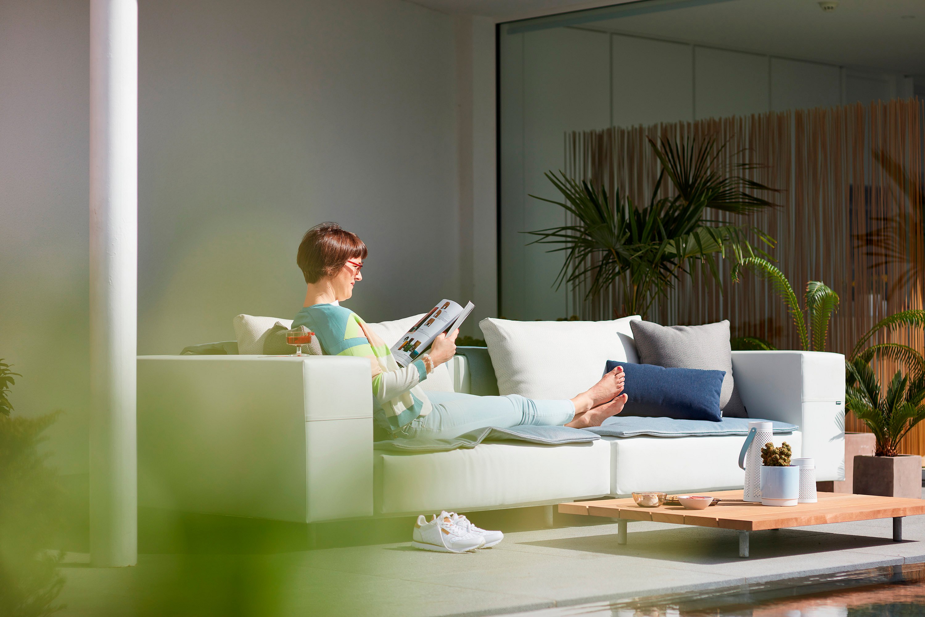   Eine Frau entspannt auf einem weißen Walrus-Sofa neben einem niedrigen Holztisch auf einer hellen, überdachten Terrasse mit Topfpflanzen.