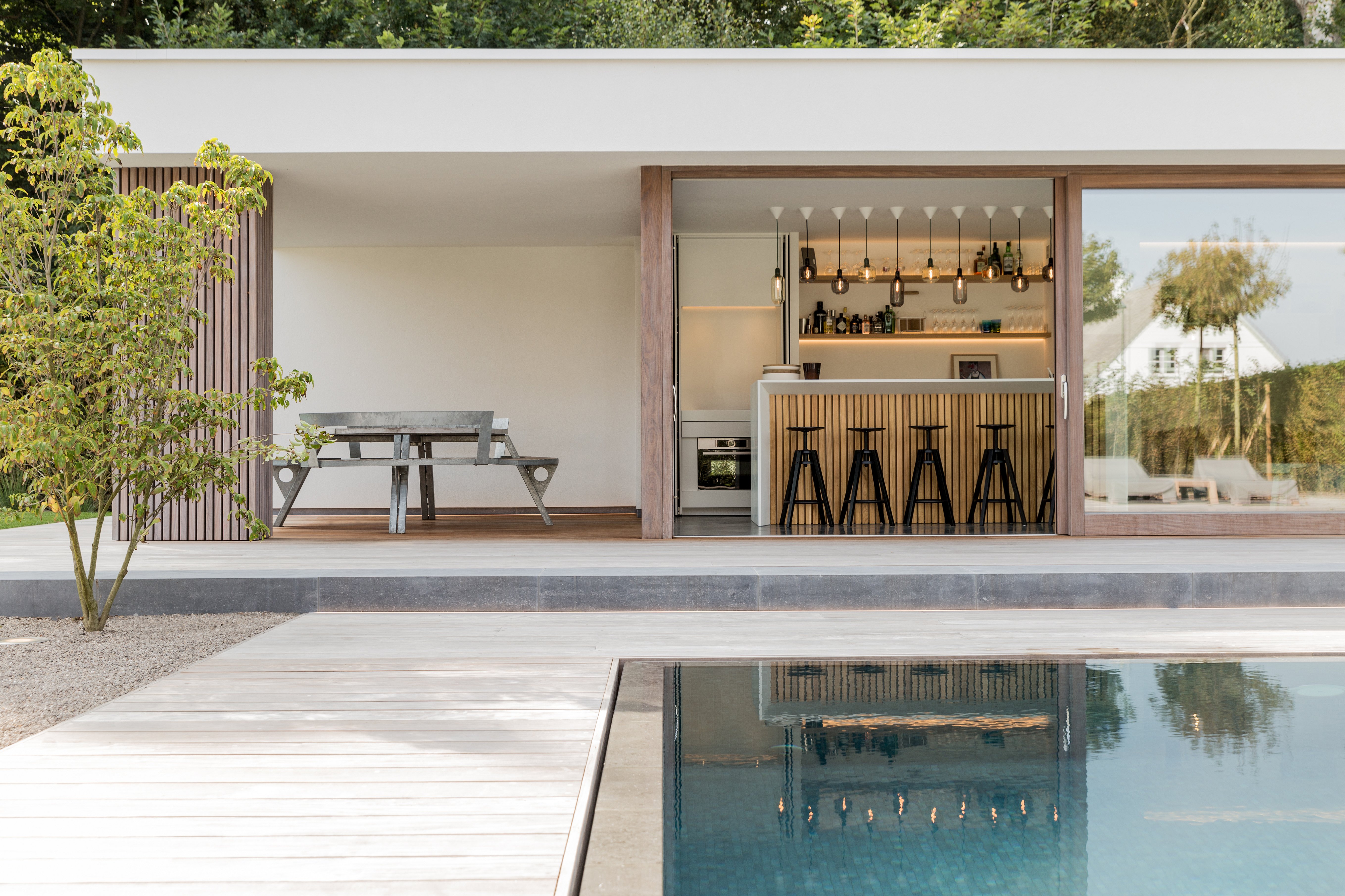 View across the swimming pool toward the poolhouse terrace, where a gargantua table sits under the roof beside a compact outdoor bar area.