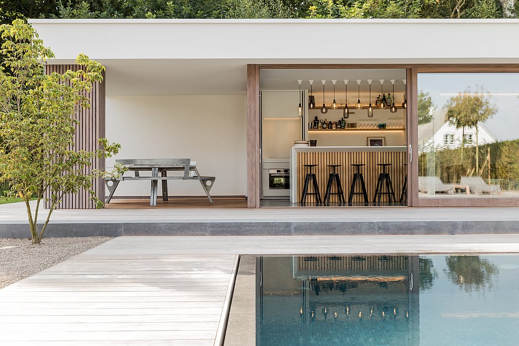 View across the swimming pool toward the poolhouse terrace, where a gargantua table sits under the roof beside a compact outdoor bar area.