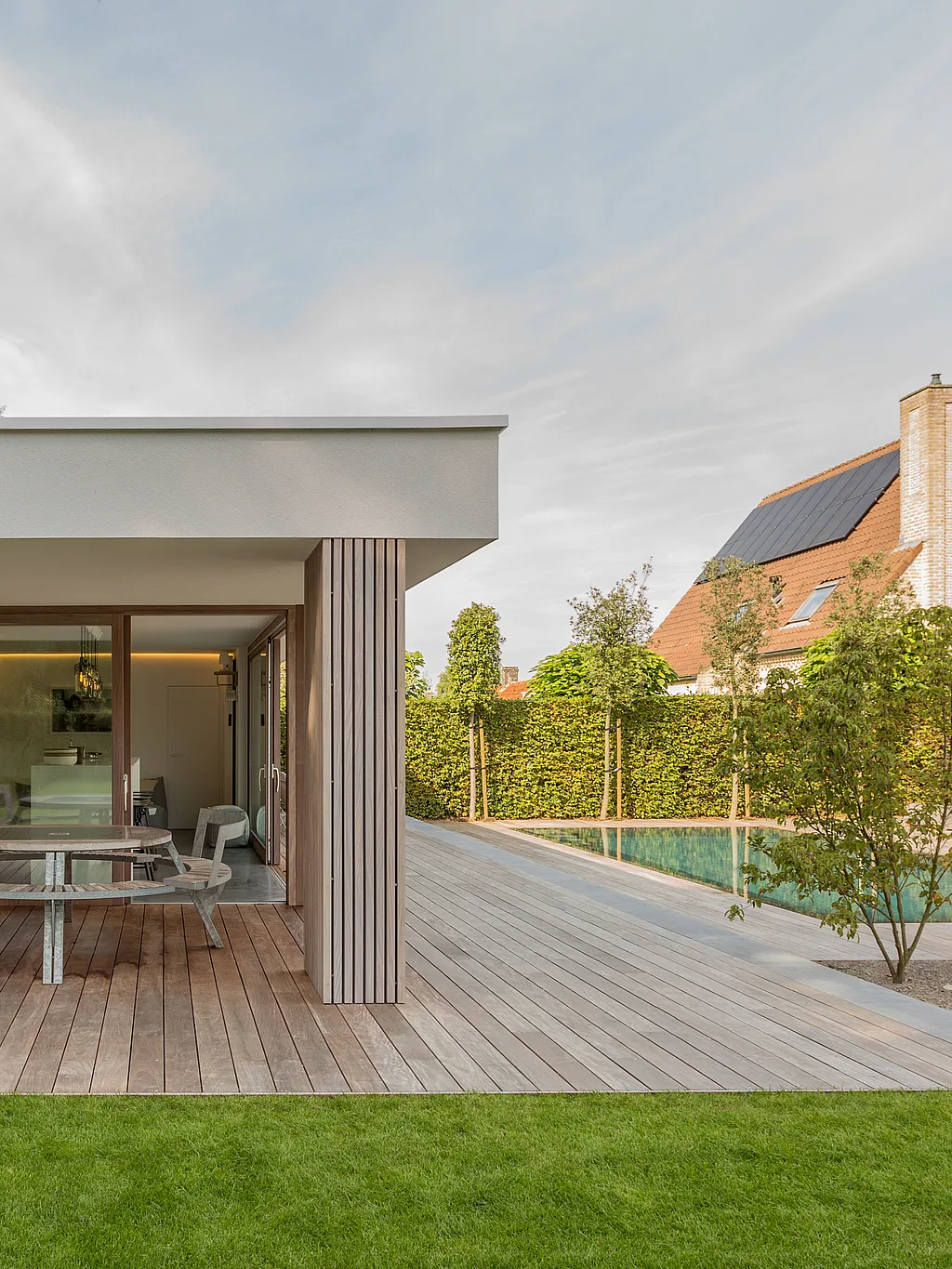 Modern wit poolhouse met een overdekt houten terras met een ronde Gargantua picknicktafel, naast een lang smal zwembad en netjes aangelegde tuin.