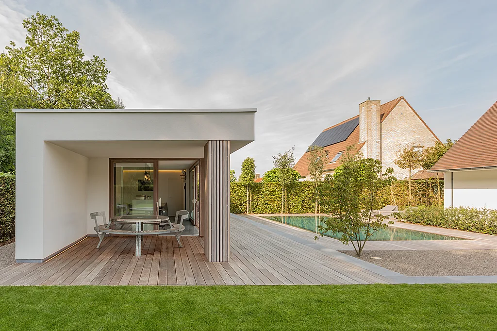 Pool house moderne blanc avec une terrasse en bois couverte dotée d’une table de pique-nique ronde Gargantua, à côté d’une longue piscine étroite et d’un jardin soigneusement paysagé.