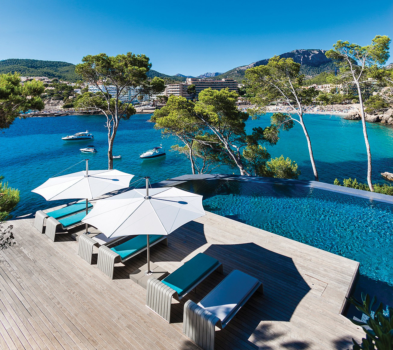 Verhoogd uitzicht op een houten pooldeck in Mallorca met witte parasols die turquoise ligstoelen overschaduwen naast een overloopzwembad, met uitzicht op een helderblauwe baai met pijnbomen, boten en bergen.