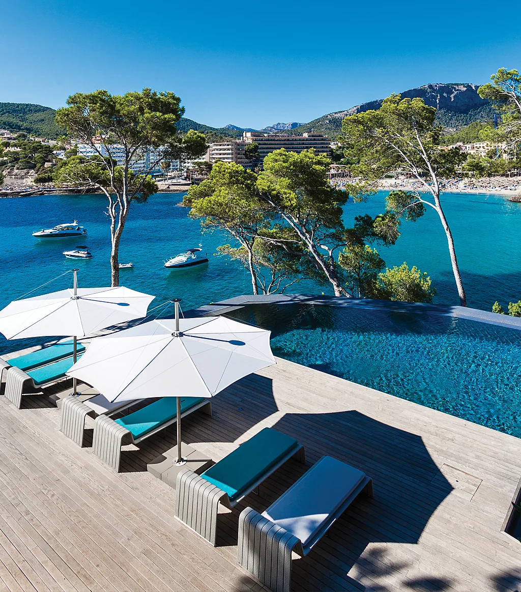 Verhoogd uitzicht op een houten pooldeck in Mallorca met witte parasols die turquoise ligstoelen overschaduwen naast een overloopzwembad, met uitzicht op een helderblauwe baai met pijnbomen, boten en bergen.