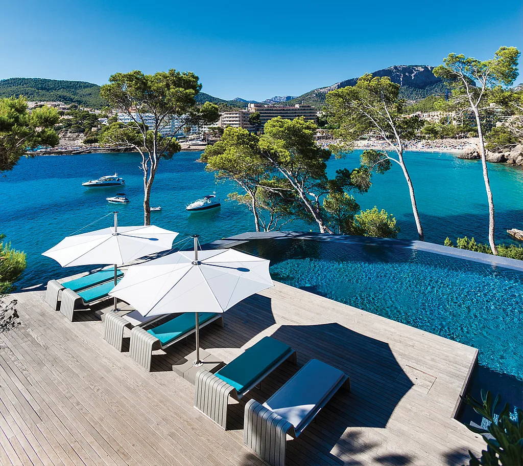 Elevated view of a wooden pool deck in Mallorca with white parasols shading turquoise sun loungers beside an infinity pool, overlooking a bright blue bay with pine trees, boats, and mountains.
