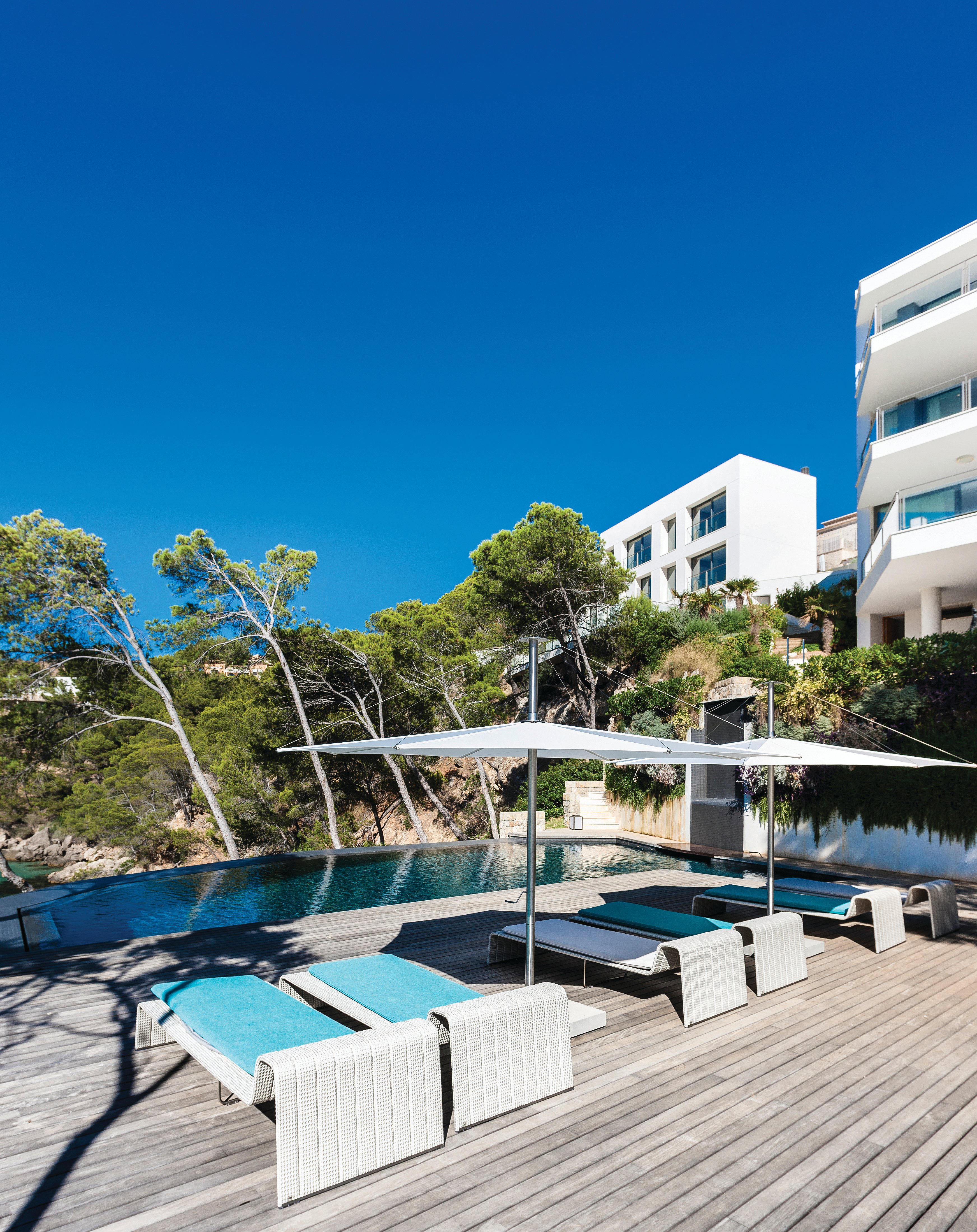 Pooldeck aus Holz mit weißen Sonnenschirmen und türkisfarbenen Liegen neben einem Infinity-Pool, im Hintergrund weiße, moderne Gebäude und üppige Bepflanzung am Hang.