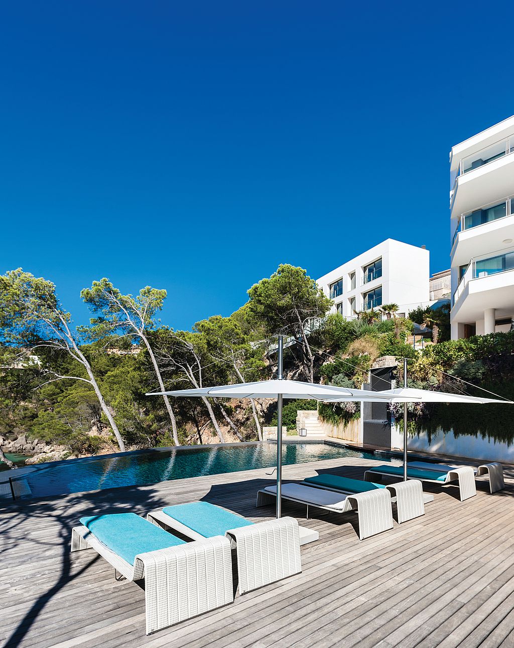 Pooldeck aus Holz mit weißen Sonnenschirmen und türkisfarbenen Liegen neben einem Infinity-Pool, im Hintergrund weiße, moderne Gebäude und üppige Bepflanzung am Hang.