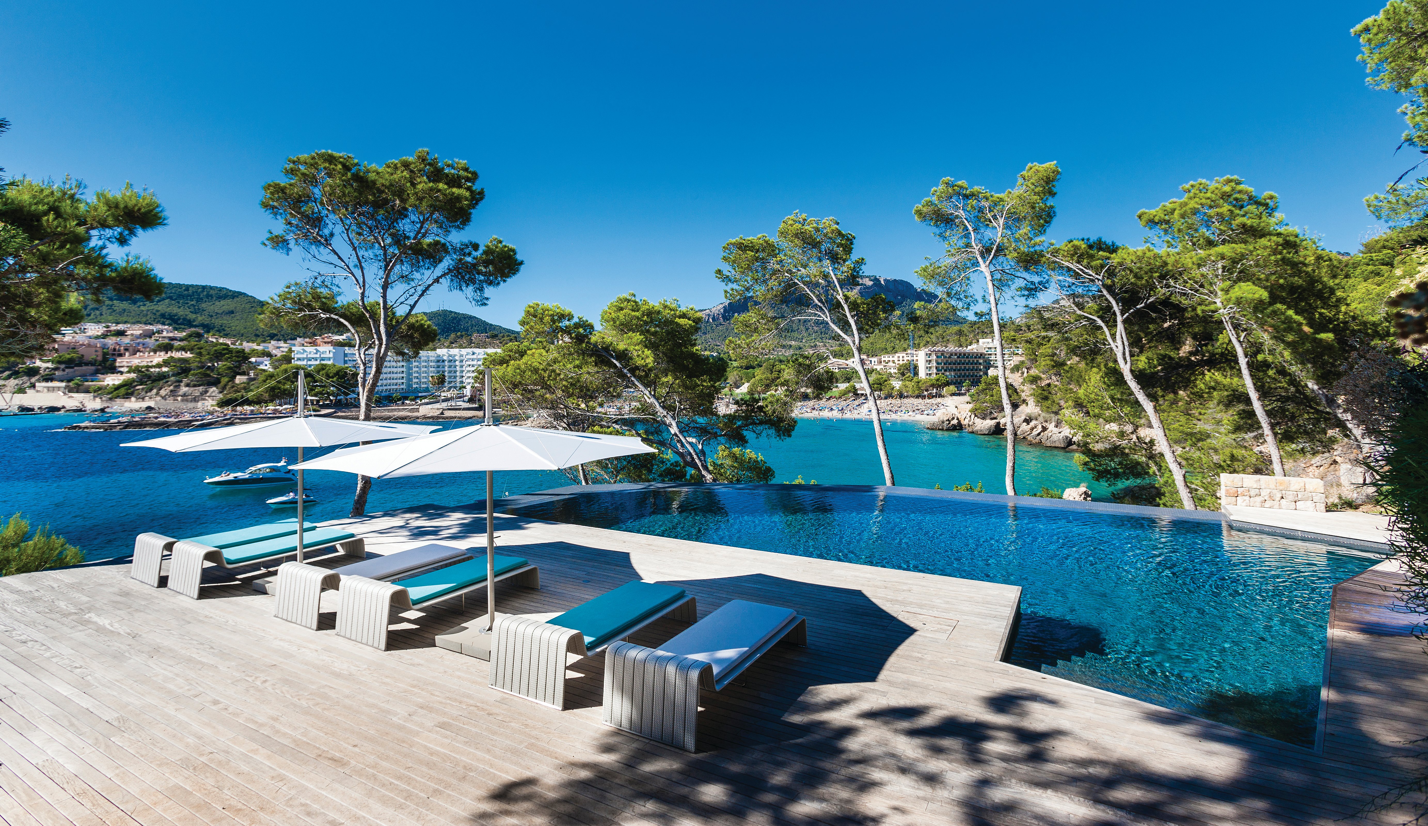 Weite Aussicht auf weiße Inumbra-Sonnenschirme und türkisfarbene Liegen, die auf einem Holzdeck neben einem Infinity-Pool aufgereiht sind, mit einer felsigen Bucht, Kiefern und Gebäuden am Meer im Hintergrund.