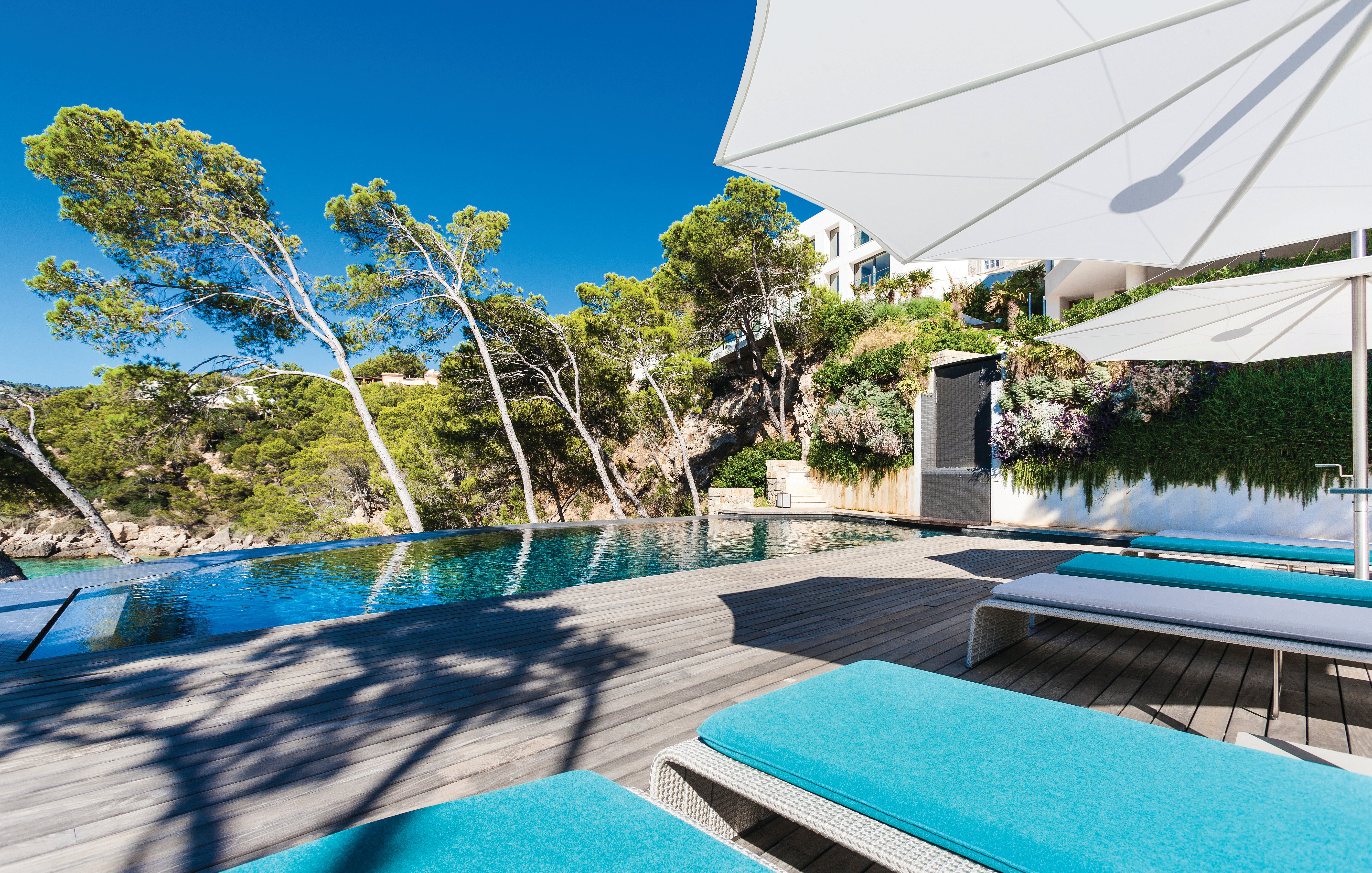 Poolterrasse mit türkis gepolsterten Liegen unter großen weißen Inumbra-Sonnenschirmen neben einem Infinity-Pool, eingerahmt von schiefen Kiefern und einem bepflanzten Hang.
