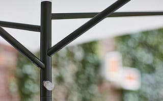 Close-up van de zwarte centrale paal en schuine steunarmen van een compacte parasol, met een zacht gefocuste bakstenen muur en klimop op de achtergrond.