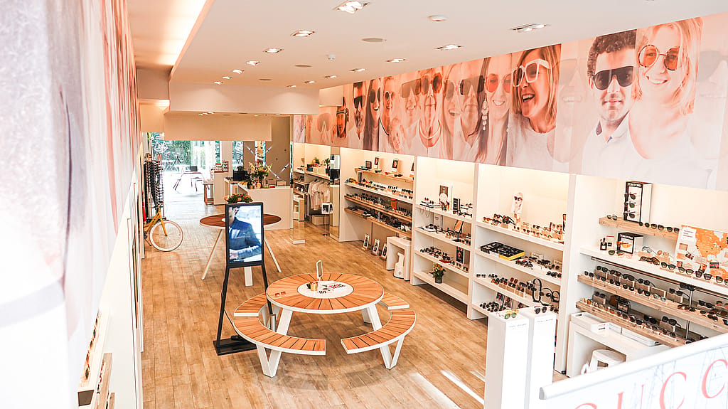 Helder interieur van de brillenwinkel met witte displayplanken, een digitaal scherm, fietsen en ronde houten tafels met een pantagruel zitbanken langs het middenpad.