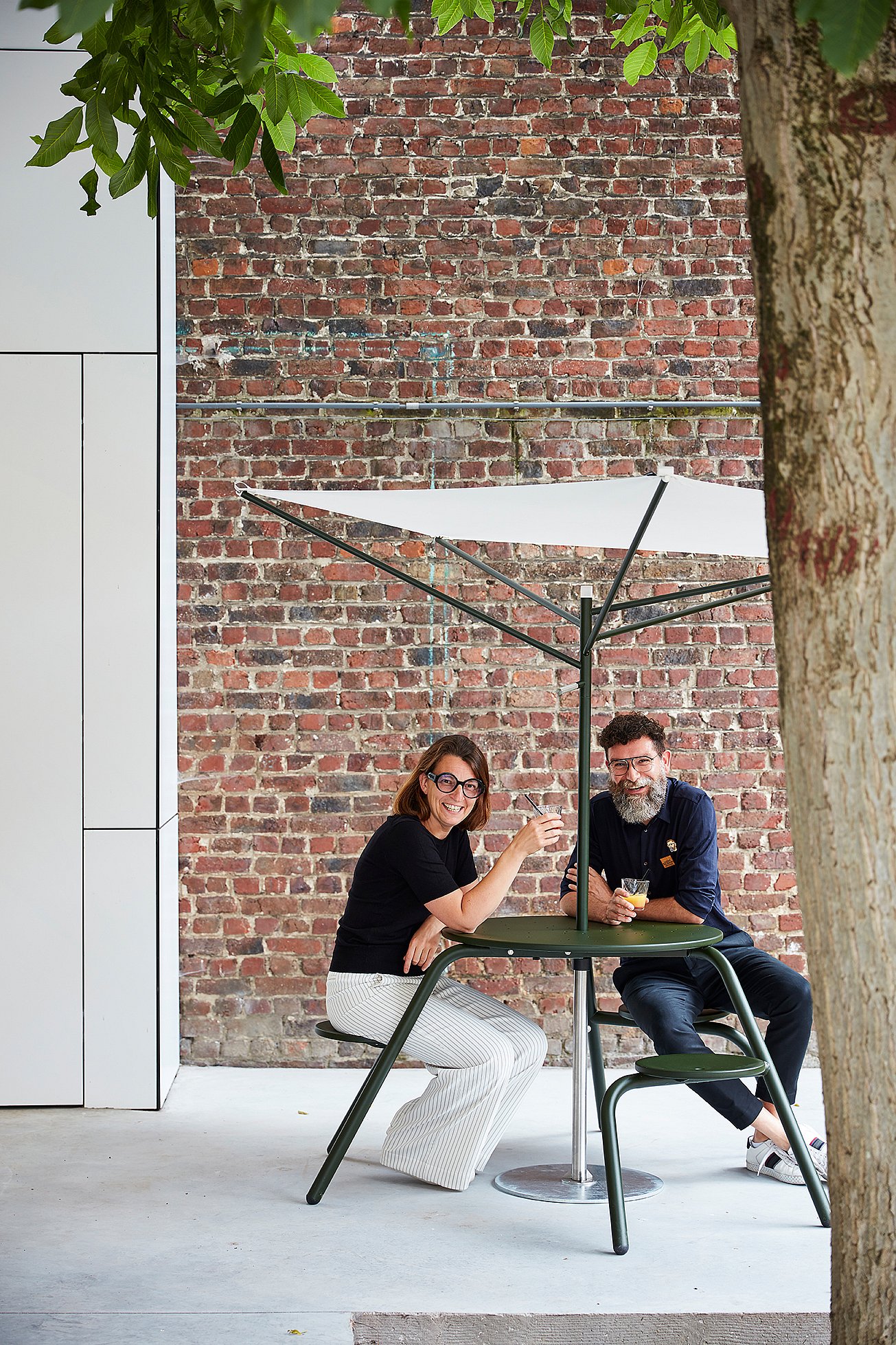 Twee mensen zitten aan een donkergroene hoge virustafel voor buiten met geïntegreerde stoelen onder een kleine witte acacia-parasol, tegen een rode bakstenen muur geplaatst en omlijst door boombladeren.
