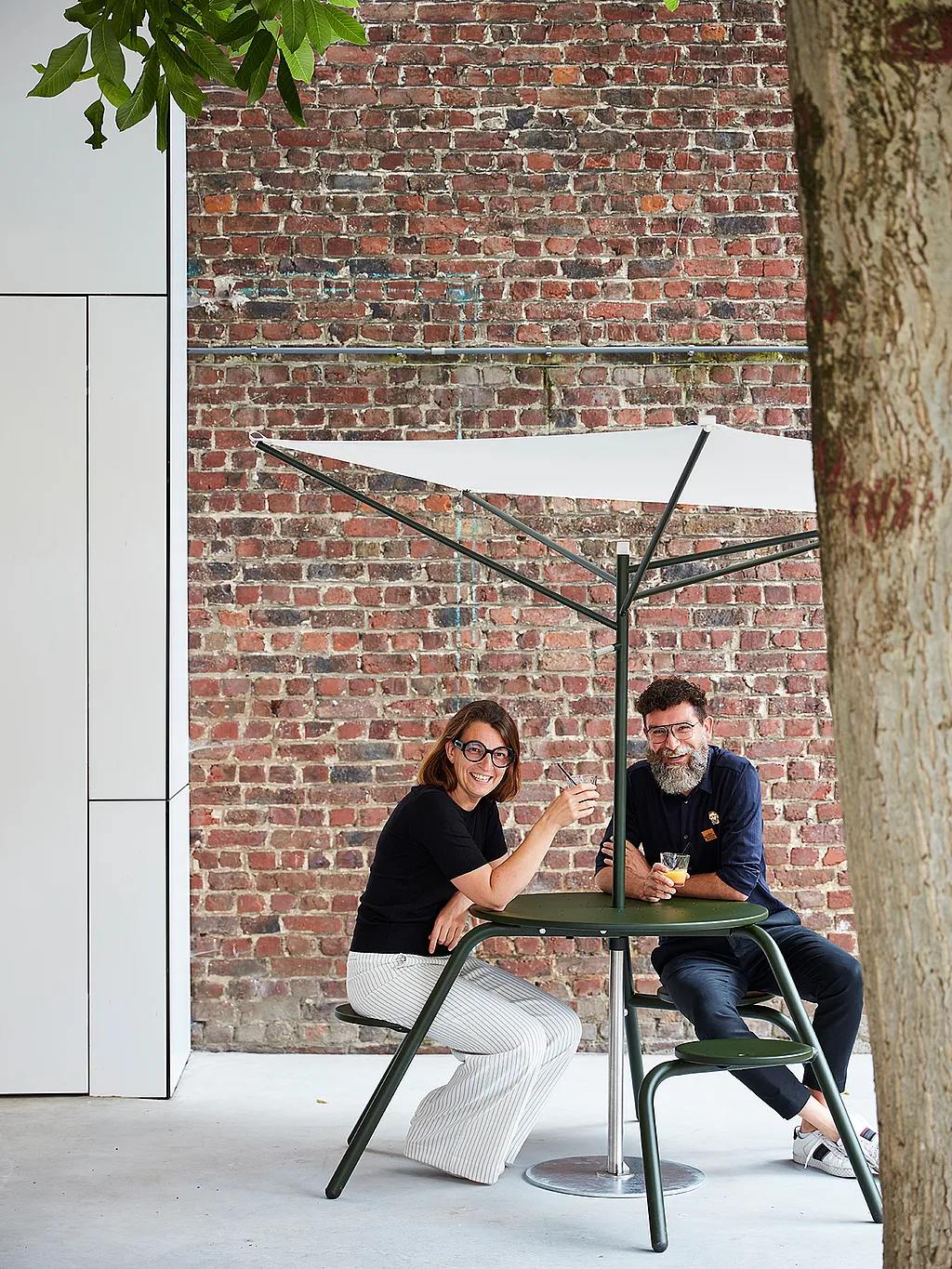 Twee mensen zitten aan een donkergroene hoge virustafel voor buiten met geïntegreerde stoelen onder een kleine witte acacia-parasol, tegen een rode bakstenen muur geplaatst en omlijst door boombladeren.