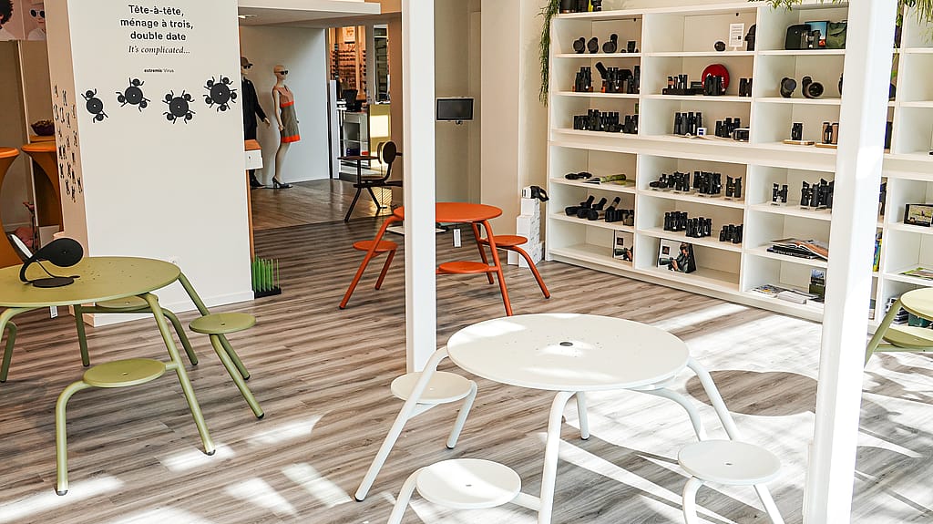 Binnenruimte met ronde virustafels in wit, saliegroen en oranje, omgeven door geïntegreerde stoelen, met wandplanken met verrekijker en andere optische apparatuur.