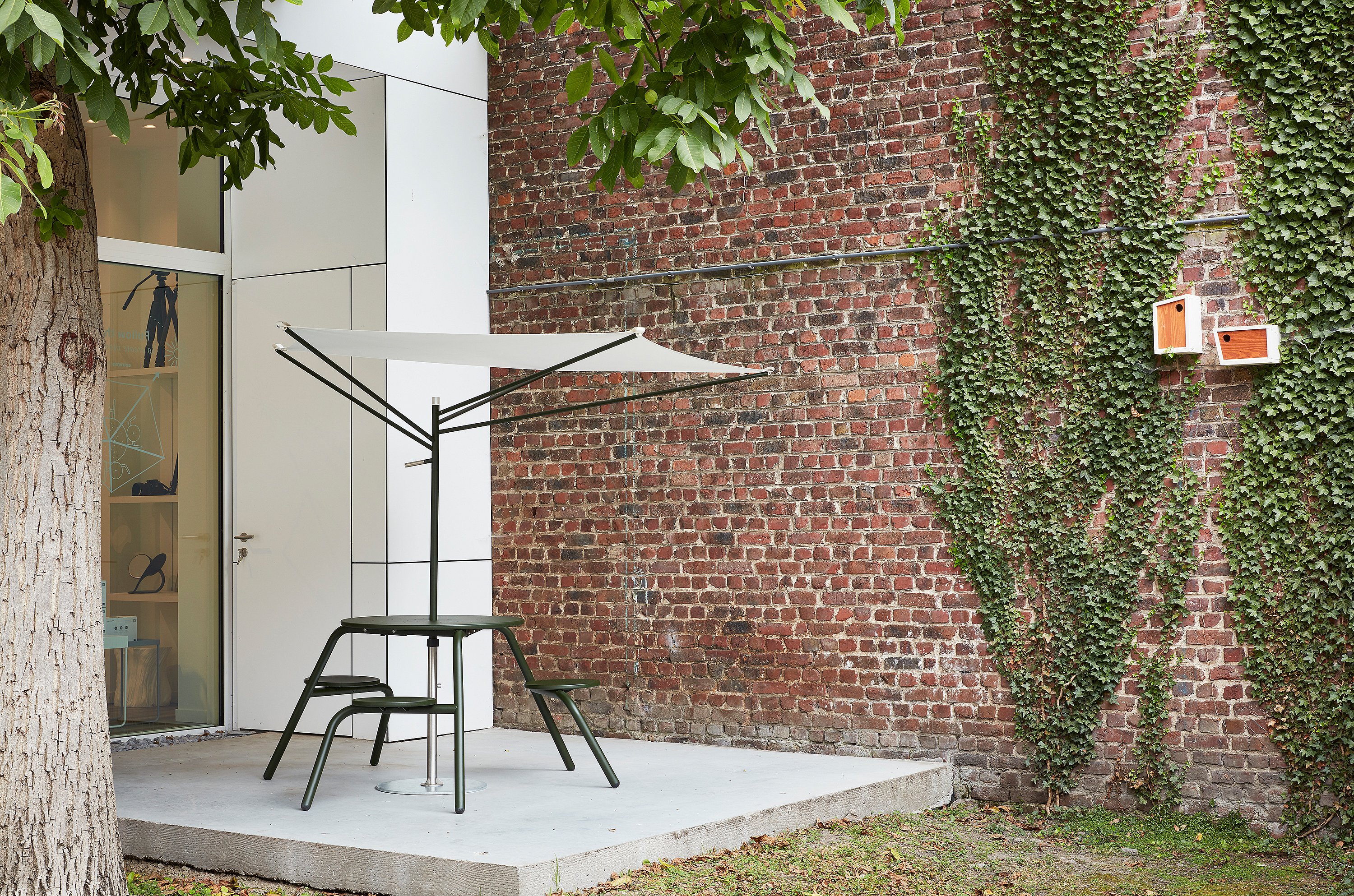 Donkergroene hoge virustafel met geïntegreerde stoelen en een compacte witte acaciaparasol op een betonnen terras naast een bakstenen muur gedeeltelijk bedekt met klimop.