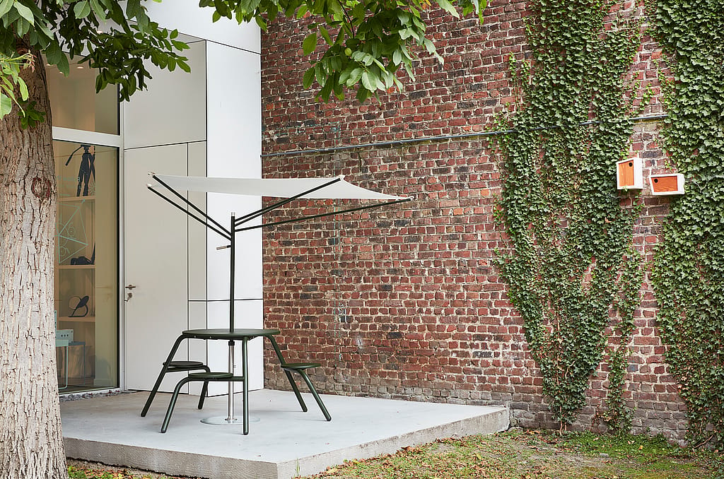 Donkergroene hoge virustafel met geïntegreerde stoelen en een compacte witte acaciaparasol op een betonnen terras naast een bakstenen muur gedeeltelijk bedekt met klimop.