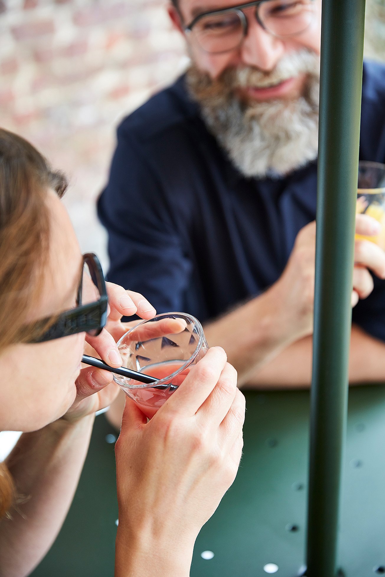 Close-up van een vrouw die een roze drankje door een rietje drinkt terwijl een bebaarde man over de tafel lacht met een glas