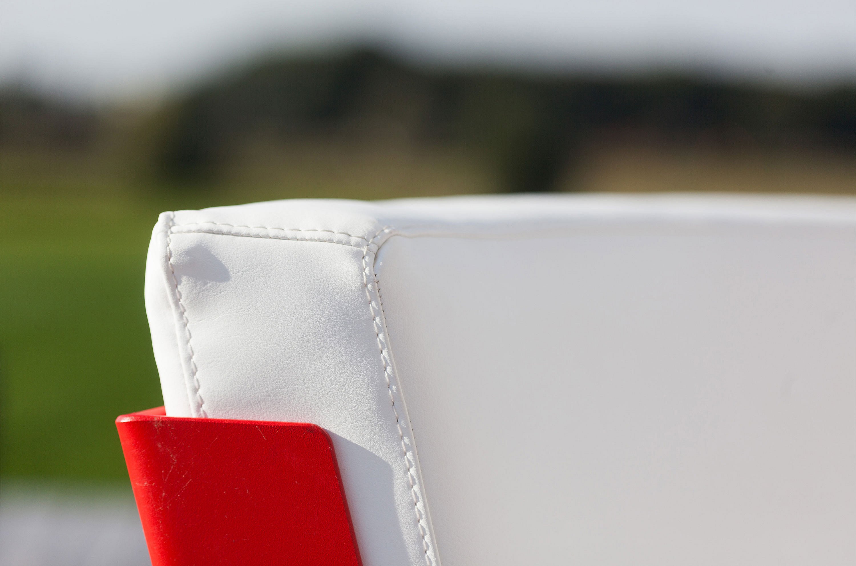 Close-up van een wit gestoffeerde rugleuninghoek met zichtbare stiksels en een rood buitenpaneel tegen een zacht wazig landschap.