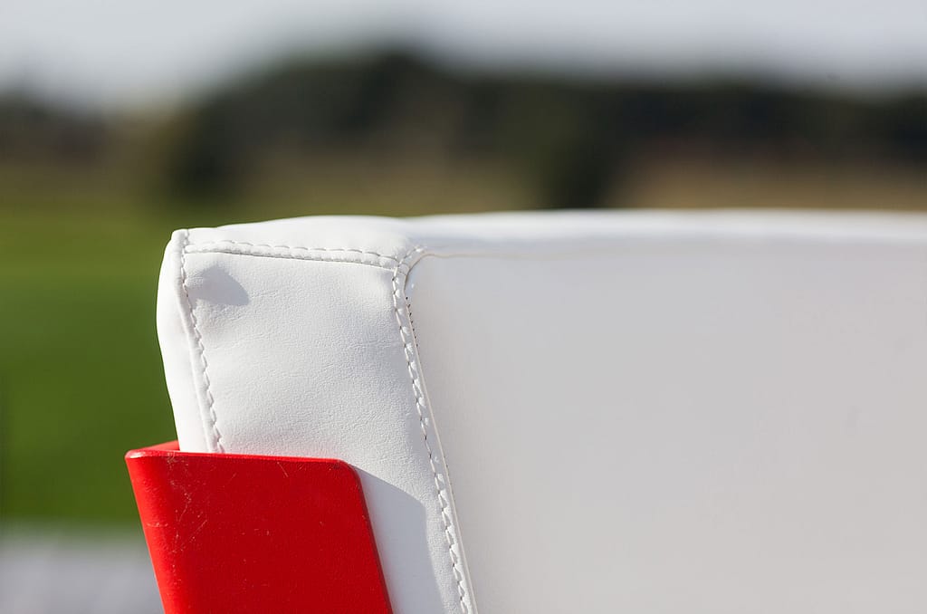 Close-up van een wit gestoffeerde rugleuninghoek met zichtbare stiksels en een rood buitenpaneel tegen een zacht wazig landschap.
