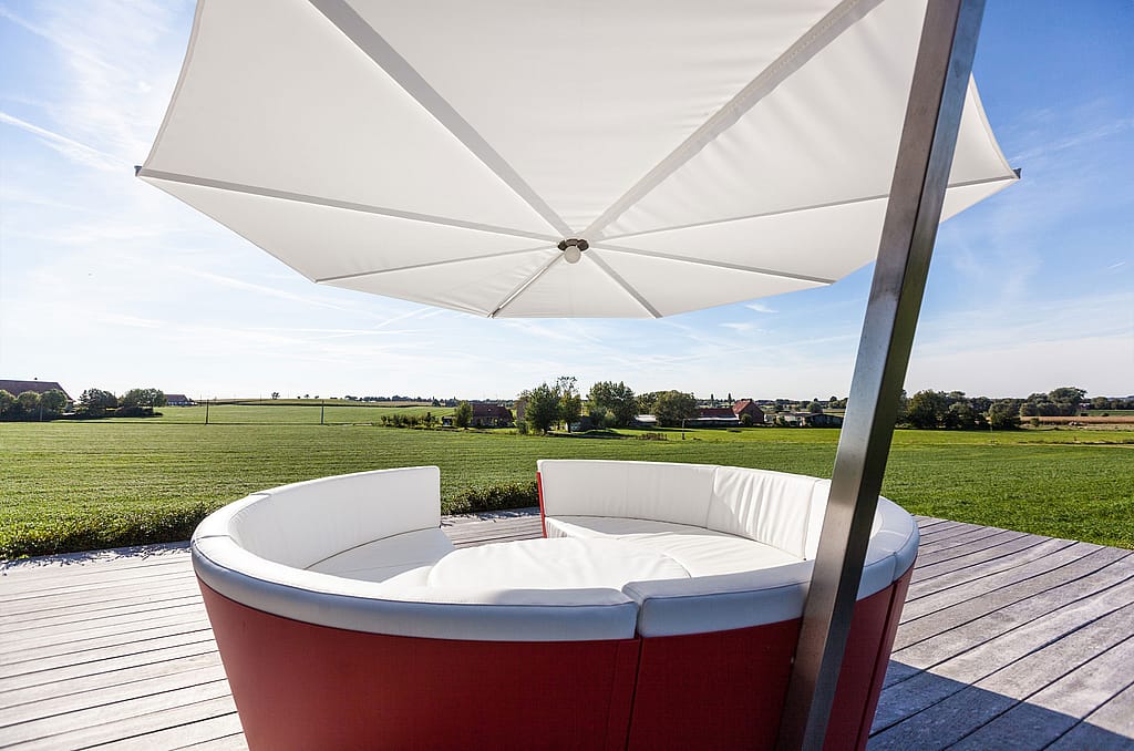 Gebogen rode kosmoslounge met witte gestoffeerde stoelen en een ronde witte tafel onder een grote witte parasol, op een houten terras met groene velden daarachter.
