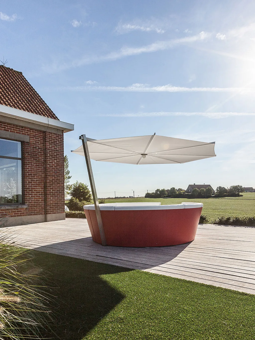 Red kosmos with white cushions and a large white parasol on a wooden terrace beside a red brick house, overlooking open countryside fields.