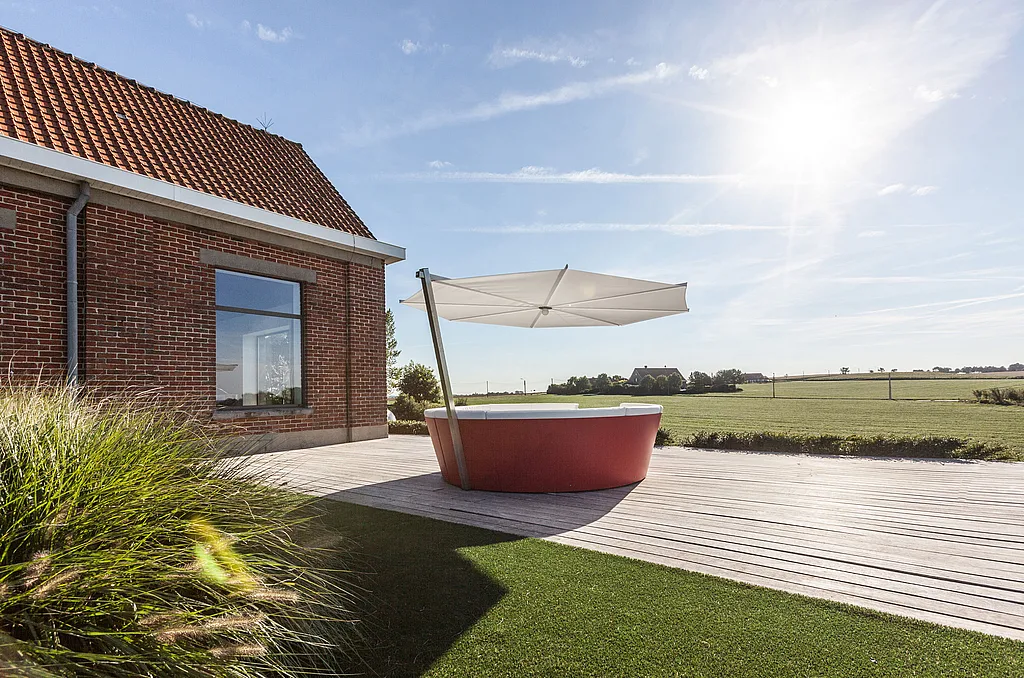 Red kosmos with white cushions and a large white parasol on a wooden terrace beside a red brick house, overlooking open countryside fields.
