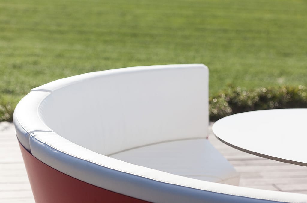Close-up van de gebogen witte rugleuning en zitkussen van de kosmoslounge buiten naast een ronde witte tafel, met een wazig groen veld erachter.
