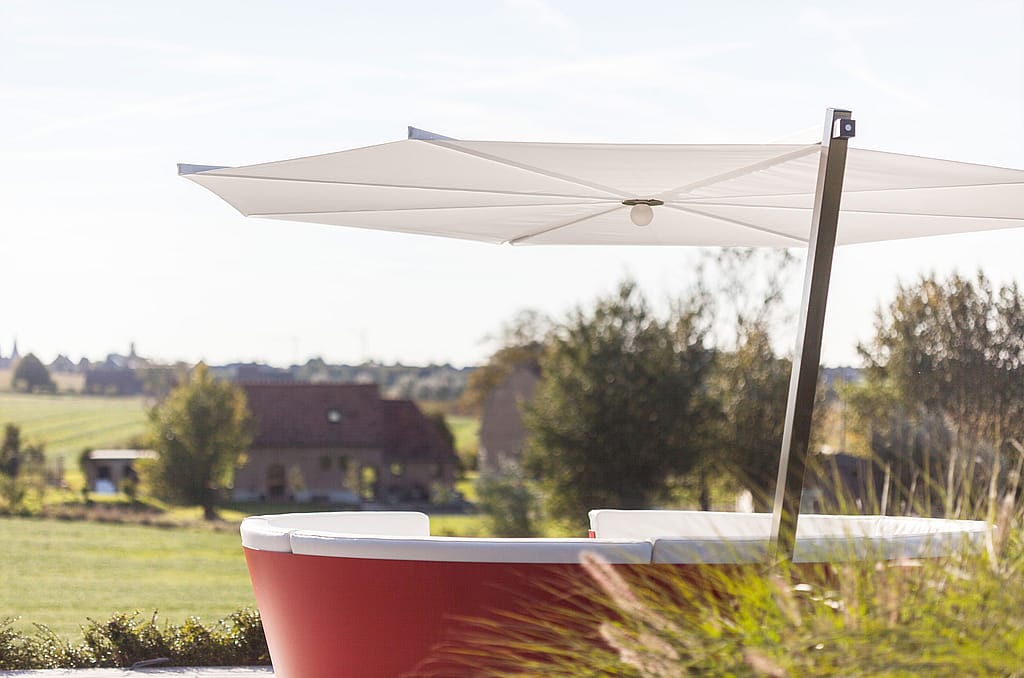 Zijaanzicht van de rode ronde kosmoslounge onder een witte parasol, met siergrassen op de voorgrond en landelijke huizen op de achtergrond.