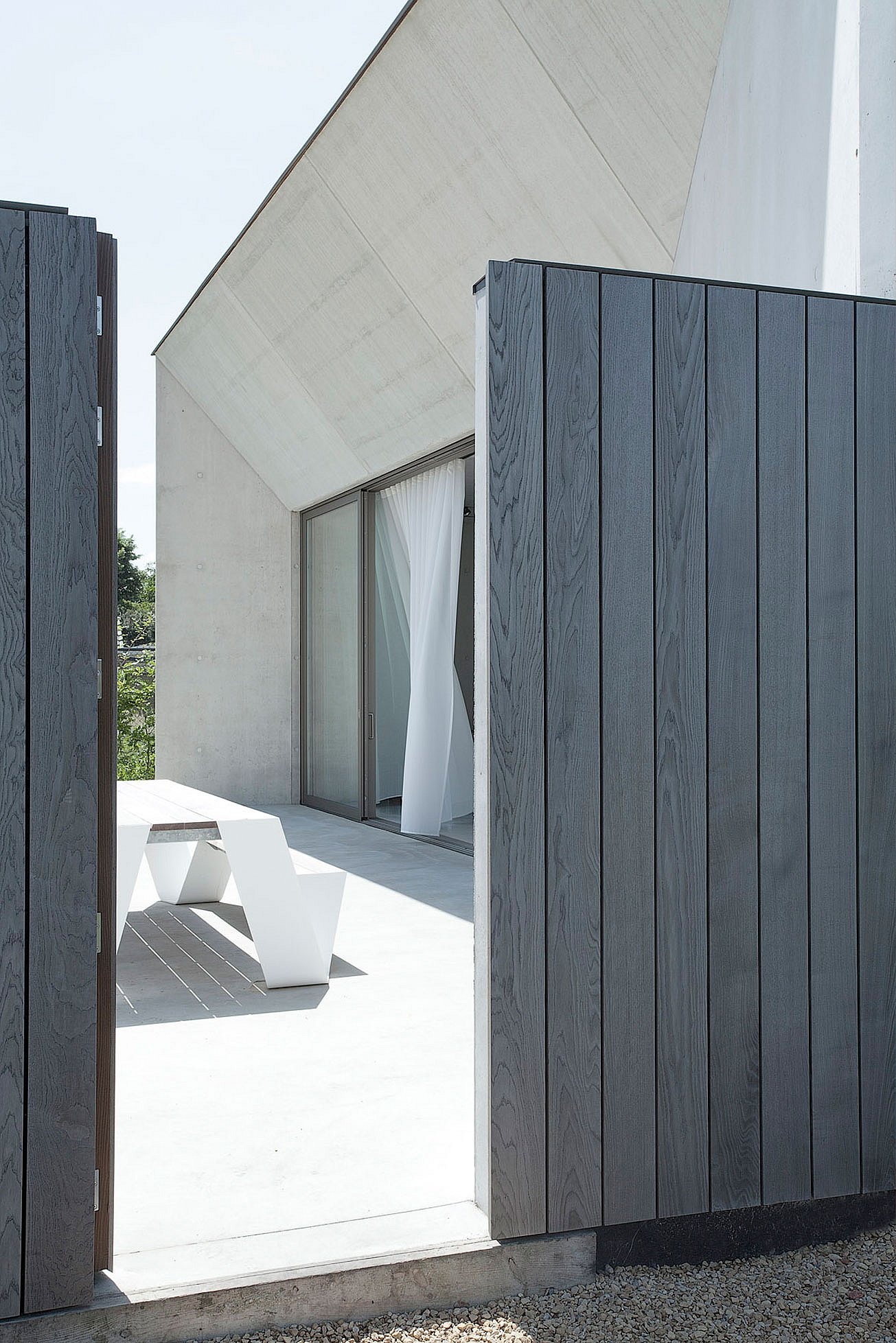 View through an open dark wooden gate toward a minimalist concrete house with a sloped roof and a white hopper picnic table on the sunlit terrace.