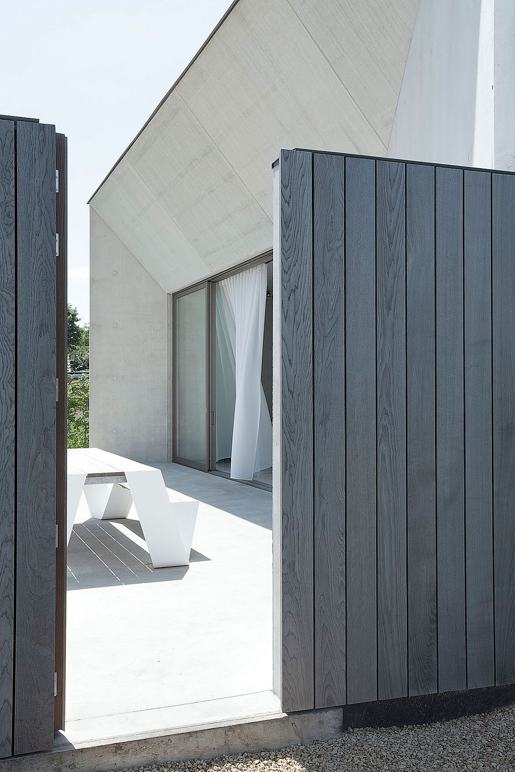 View through an open dark wooden gate toward a minimalist concrete house with a sloped roof and a white hopper picnic table on the sunlit terrace.