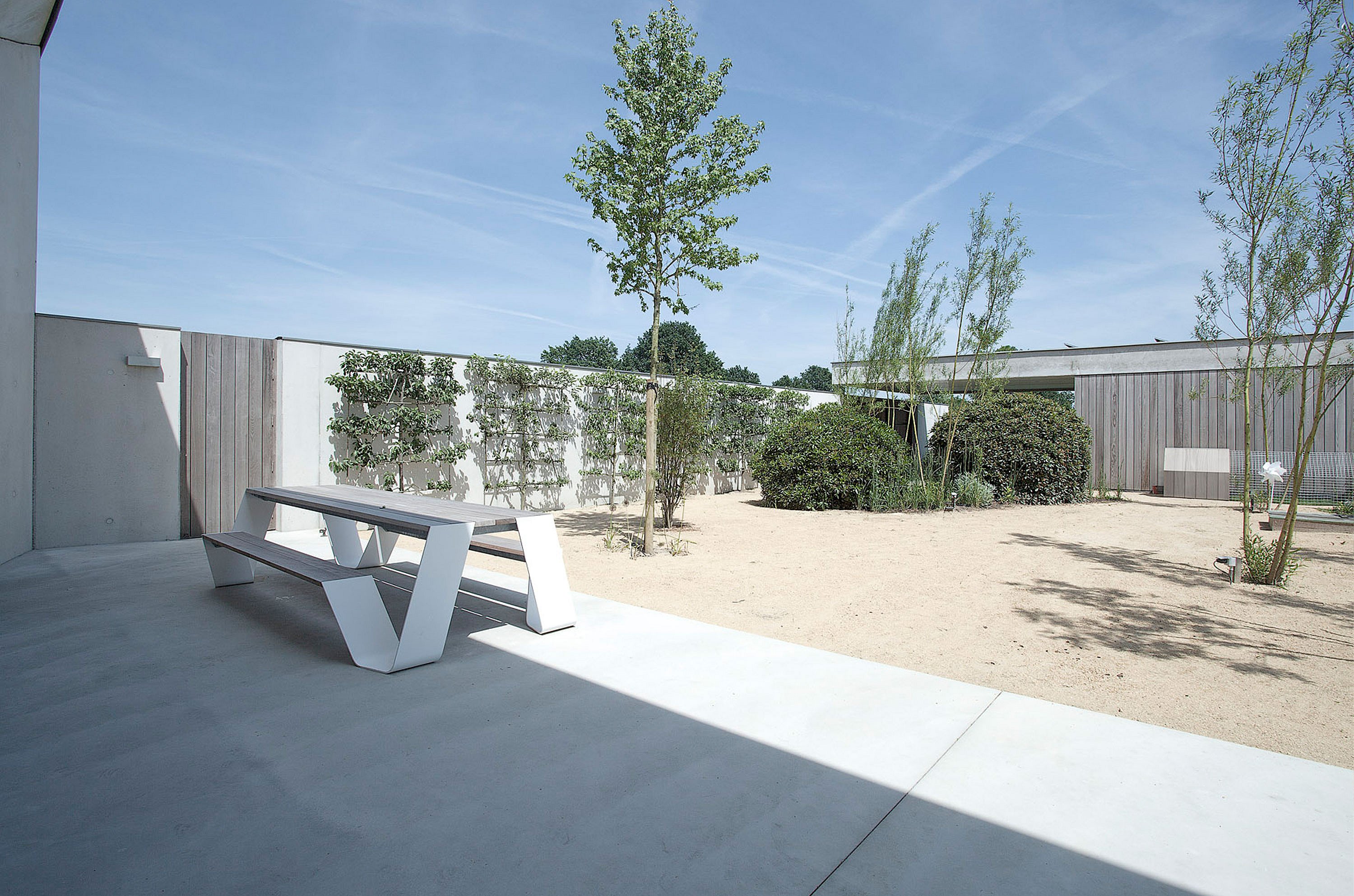 White hopper picnic table with attached bench on a shaded concrete terrace facing a sandy courtyard with young trees, shrubs, white walls, and warm wood cladding