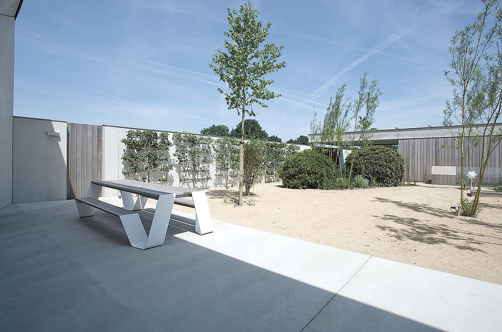 White hopper picnic table with attached bench on a shaded concrete terrace facing a sandy courtyard with young trees, shrubs, white walls, and warm wood cladding