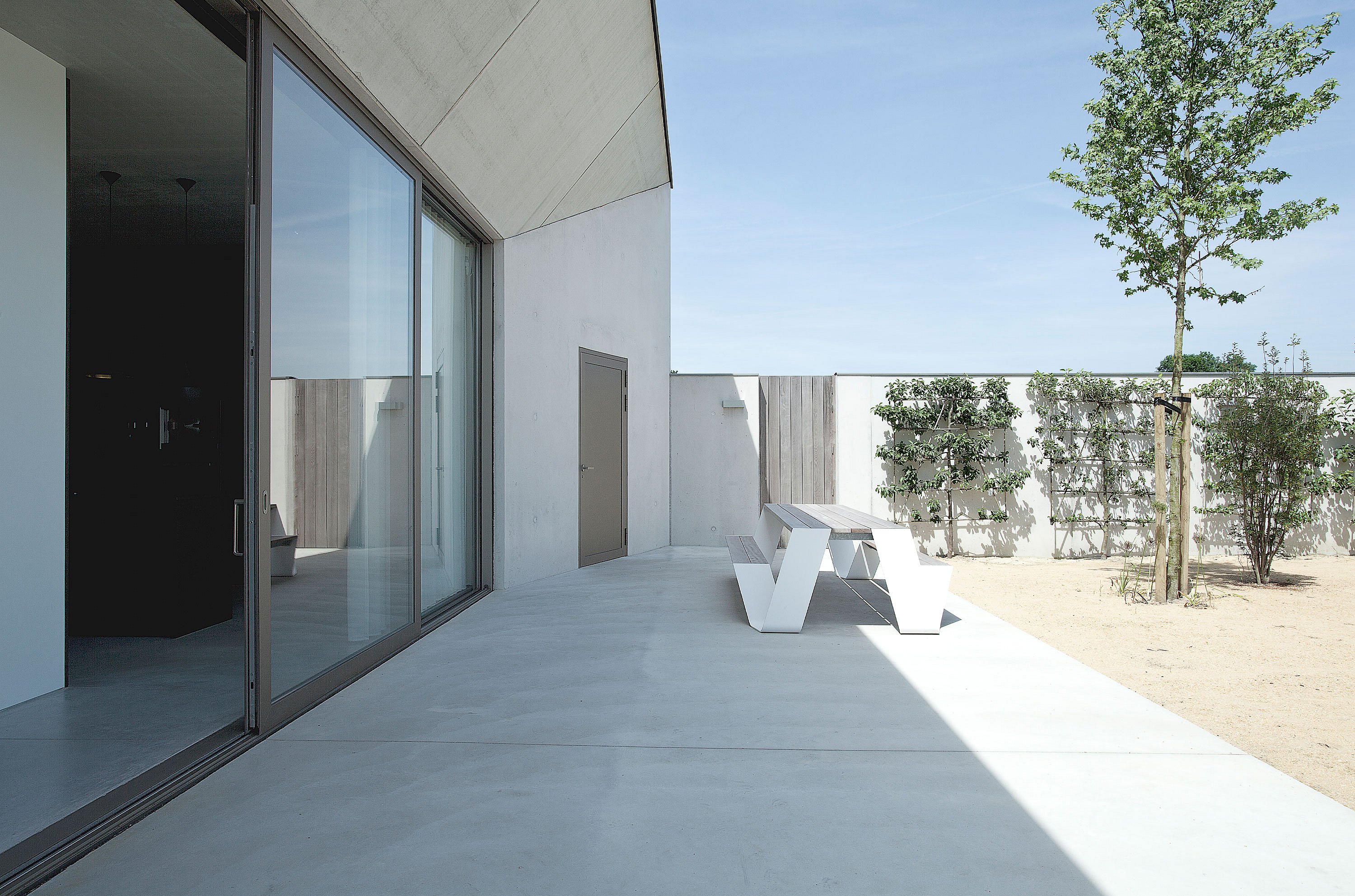 Side view along a concrete terrace with a white hopper picnic table beside floor-to-ceiling glass, overlooking a quiet courtyard with espaliered plants, shrubs, and a young tree.