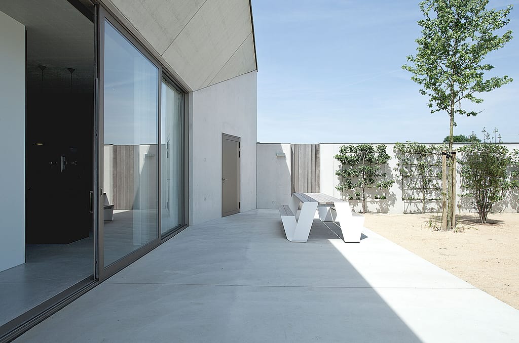 Side view along a concrete terrace with a white hopper picnic table beside floor-to-ceiling glass, overlooking a quiet courtyard with espaliered plants, shrubs, and a young tree.