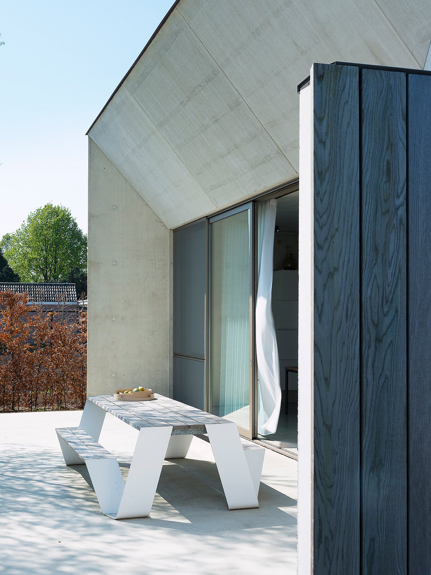 White outdoor picnic table on a pale concrete terrace beside a minimalist house with a sharply sloped concrete roof, sliding glass doors, and dark vertical wood cladding.