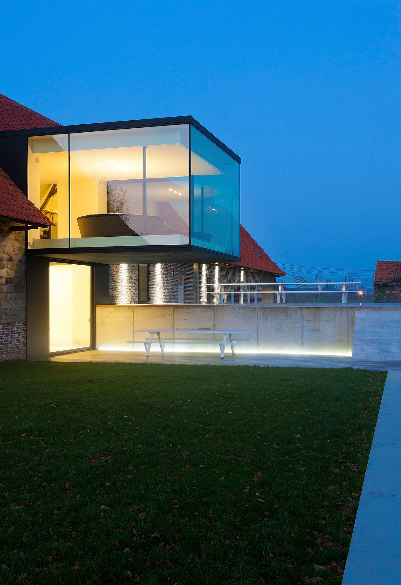 Evening exterior of the glass cube glowing with warm interior light above a lawn, with a white picnic table placed below against a concrete wall.