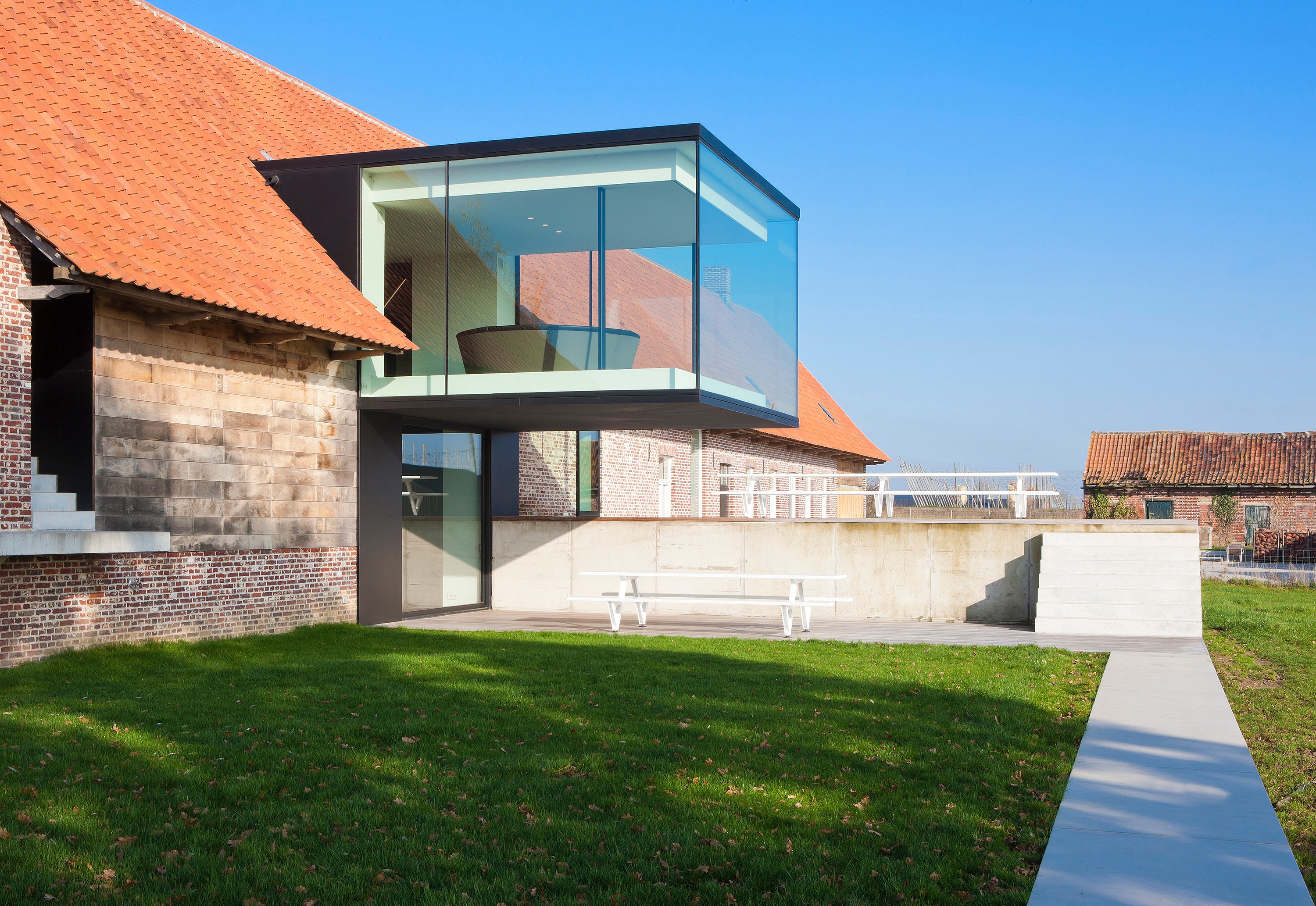 Wide exterior view of the farmhouse and projecting glass cube, with a white marina picnic table placed against a concrete wall beside a lawn.