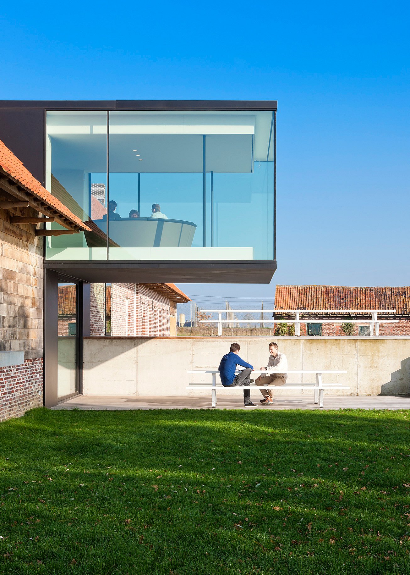 Sunlit terrace with a long white picnic table in front of a cantilevered glass cube attached to a traditional brick farmhouse with a red-tiled roof.