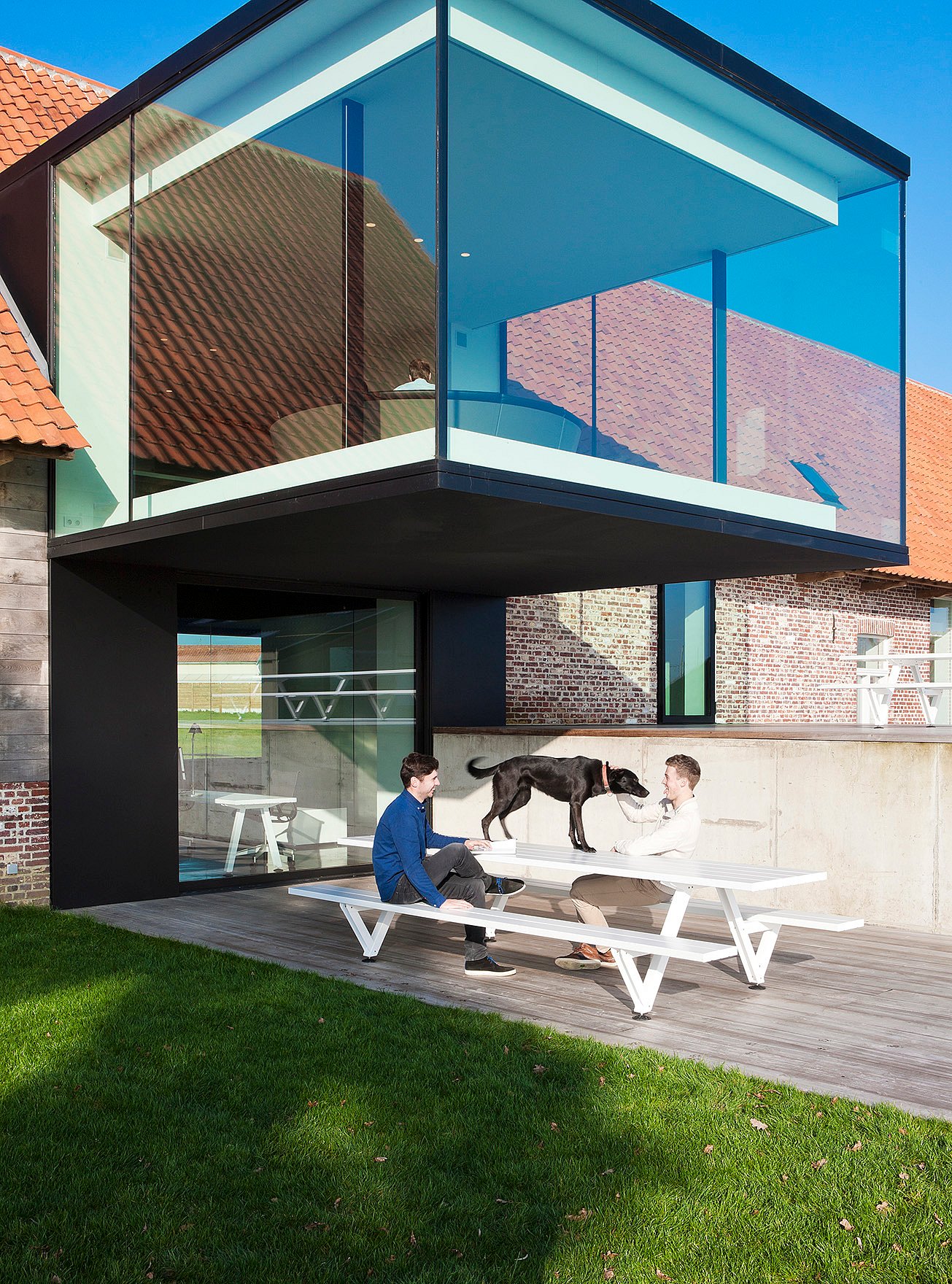 Two people seated at a white picnic table on a terrace below a glass cube, with open lawn in the foreground and people visible inside the upper room.