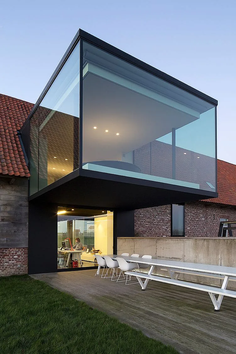 Extérieur de ferme contemporain avec un cube de mur de verre incliné au-dessus d’un pied noir, donnant sur une terrasse en bois meublée d’une longue table de pique-nique blanche et des chaises blanches du capitaine.