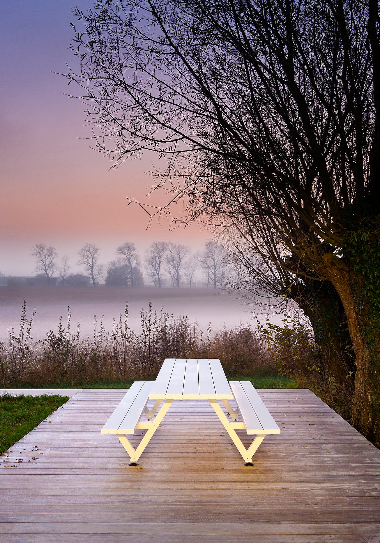 White marina picnic table on a wooden deck beside a large tree, with misty fields and a pale pink-and-lavender sky in the background.