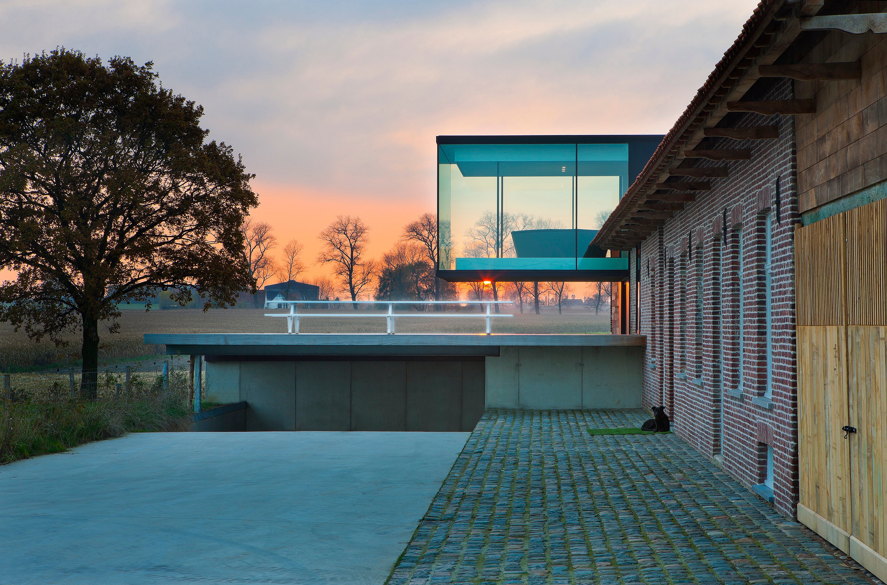 Wide dusk view of a renovated brick farmhouse with a projecting glass cube, set beside a concrete courtyard and open fields at sunset.