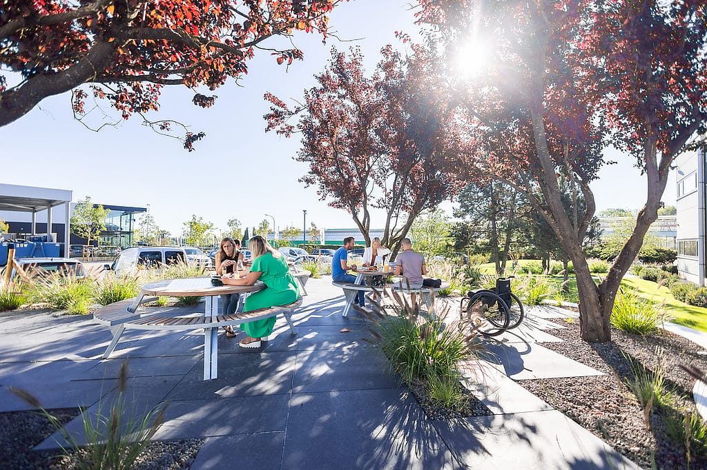 Sonnenbeschienene Gartenterrasse mit zwei runden Gargantua-Picknicktischen unter Bäumen, an denen kleine Gruppen sitzen und sich unterhalten; neben einem der Tische steht ein Fahrrad.