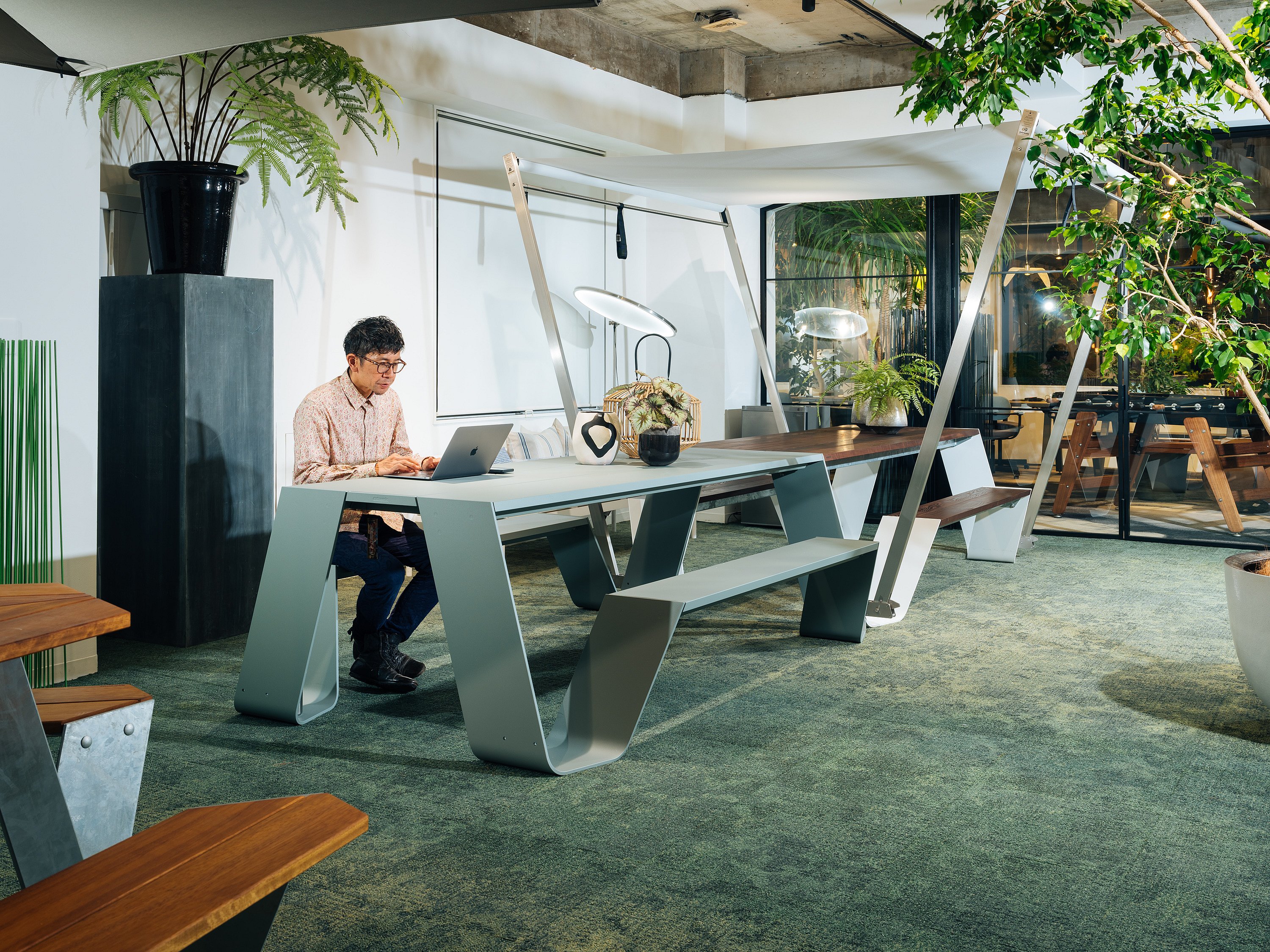   Ein Mann sitzt allein mit seinem Laptop an einem großen, hellgrauen Tisch mit Sitzbank und Baldachin in einem geräumigen, mit Pflanzen gefüllten Büro.