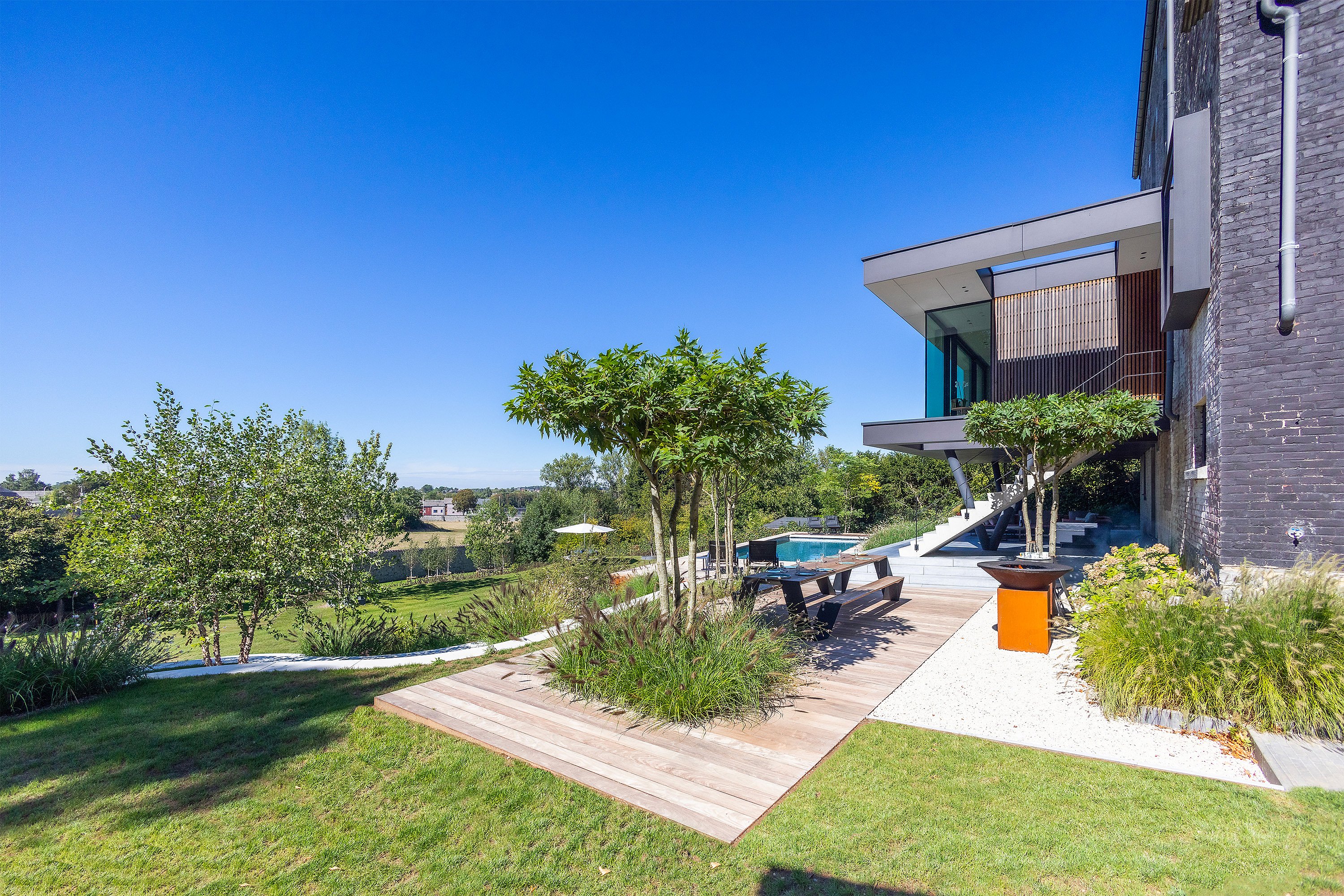 Weite Tagesansicht eines modernen Hauses und Terrassengartens an einem Hang mit geschichteter Bepflanzung, einer hölzernen Essterrasse und einem Pool neben dem Haus unter klarem blauen Himmel.