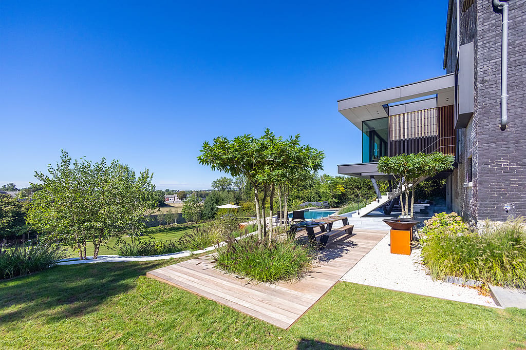Weite Tagesansicht eines modernen Hauses und Terrassengartens an einem Hang mit geschichteter Bepflanzung, einer hölzernen Essterrasse und einem Pool neben dem Haus unter klarem blauen Himmel.