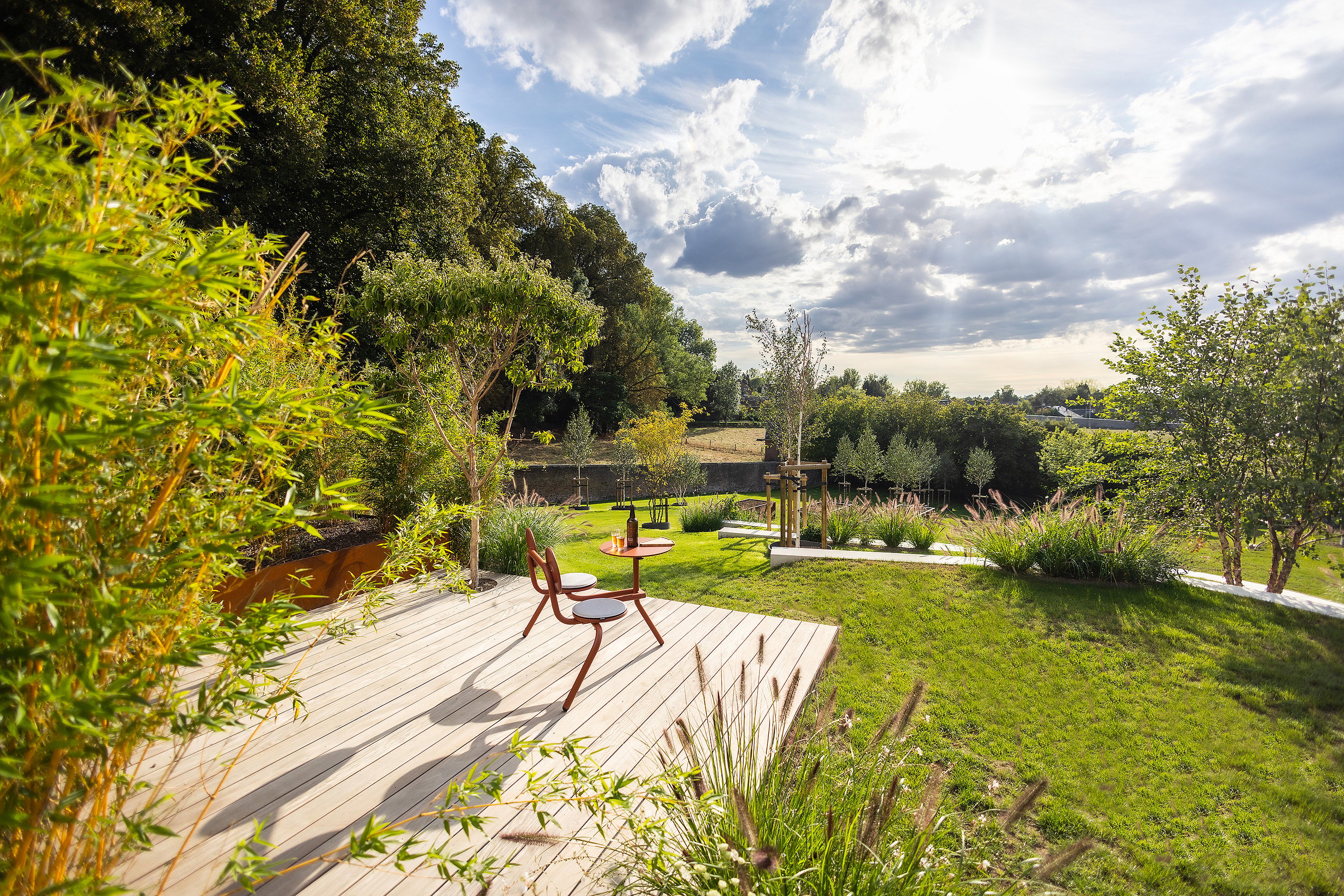 Kleiner Bistro-Zweisitzertisch auf einer Aussichtsplattform mit Flasche und Gläsern, mit Blick auf einen sonnenbeschienenen Rasen, mehrschichtige Bepflanzung und terrassierte Gartenwege