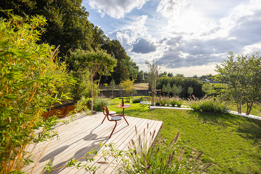 Kleiner Bistro-Zweisitzertisch auf einer Aussichtsplattform mit Flasche und Gläsern, mit Blick auf einen sonnenbeschienenen Rasen, mehrschichtige Bepflanzung und terrassierte Gartenwege