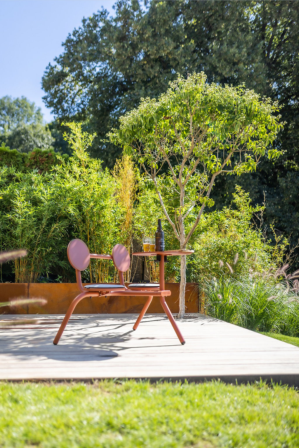 Rostfarbenes Bistro mit einem kleinen runden Tisch auf einer Holzterrasse, inmitten von dichtem Grün und Ziergräsern in einer sonnigen Gartenecke.