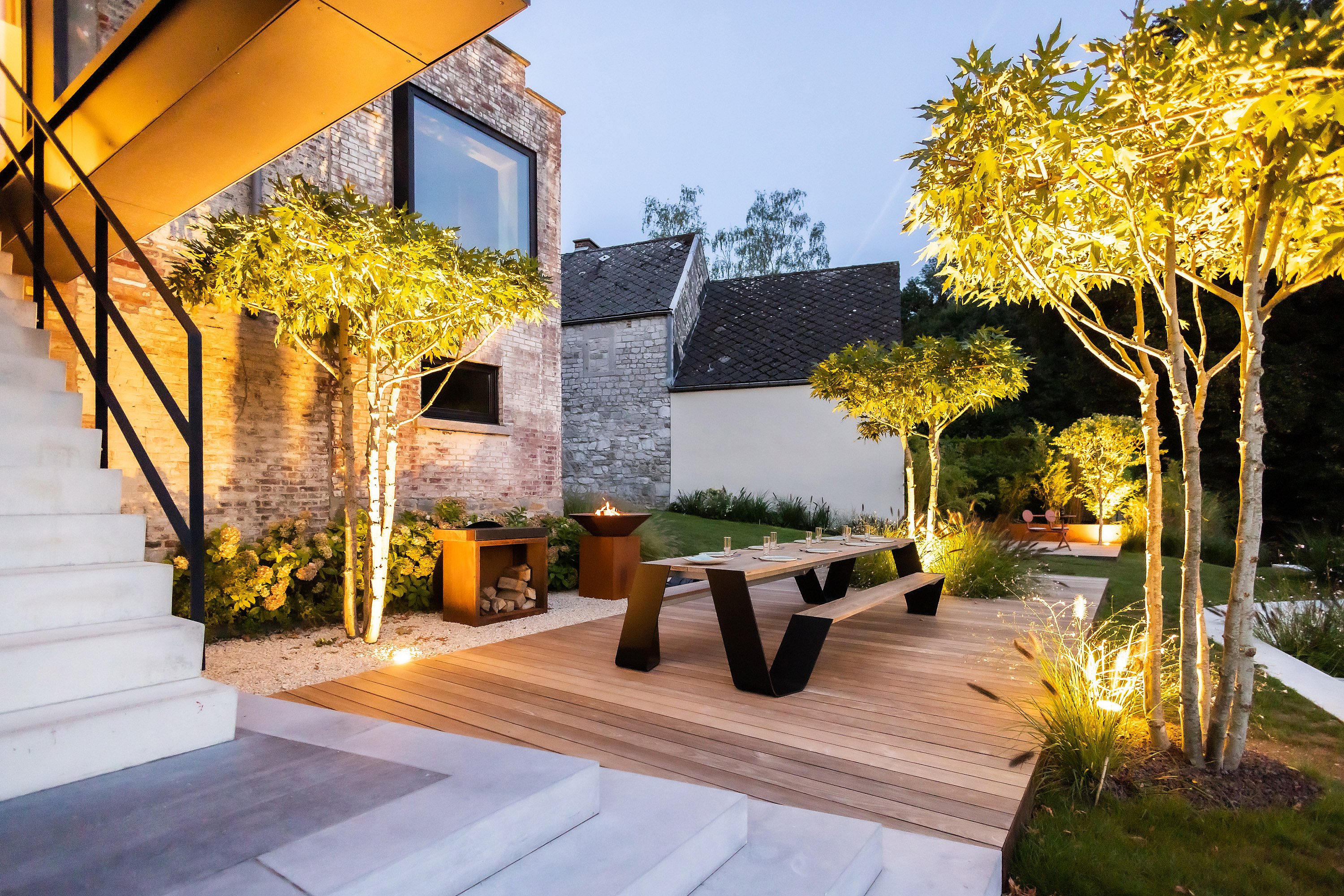 Avondzicht op een terrasvormige tuin naast een bakstenen huis, met warm verlichte gebeeldhouwde bomen, een lange houten eettafel met banken op een dek en een loungeruimte die in de verte gloeit.
